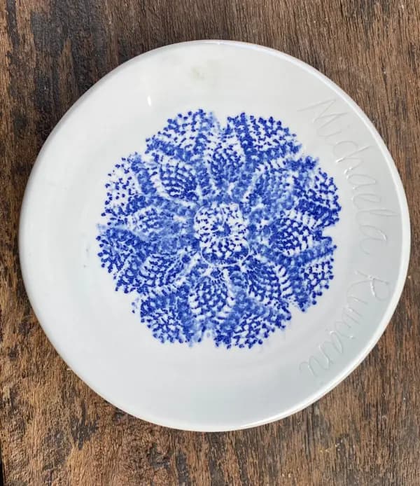 White ceramic trinket plate with a central blue lace-like pattern and embossed text on a wood background.