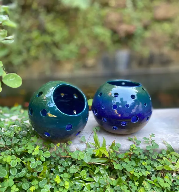 Two spherical ceramic candle holders with circular cutouts, glazed in dark green and cobalt blue, set outdoors.