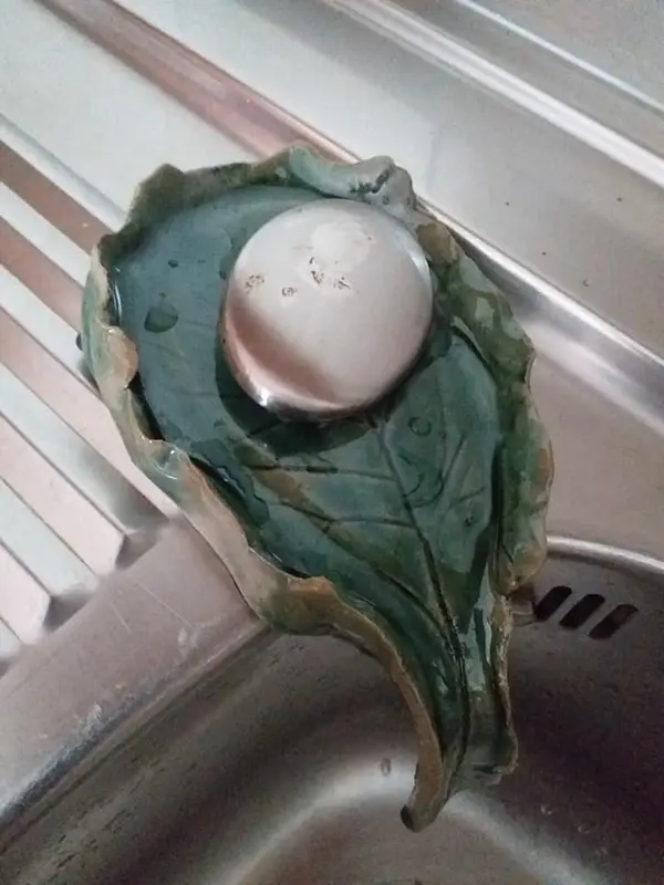 Dark green leaf-shaped ceramic soap dish holding a stainless steel soap bar on the edge of a metal sink.