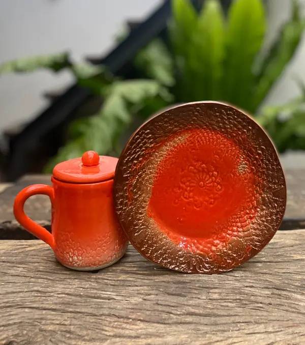 Vibrant orange lidded mug and matching textured plate with metallic bronze accents on a weathered wood surface.