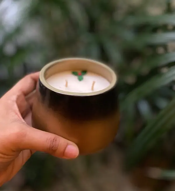 Two-tone black and beige ceramic candle holder with a double-wick holly candle inside, held against a leafy backdrop.