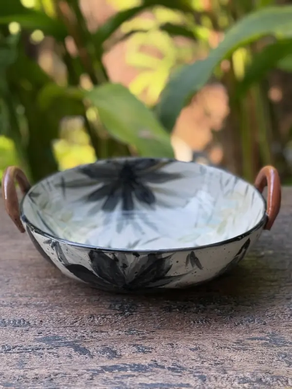 Hand-painted ceramic bowl with black floral patterns and two brown handles on a wooden surface.