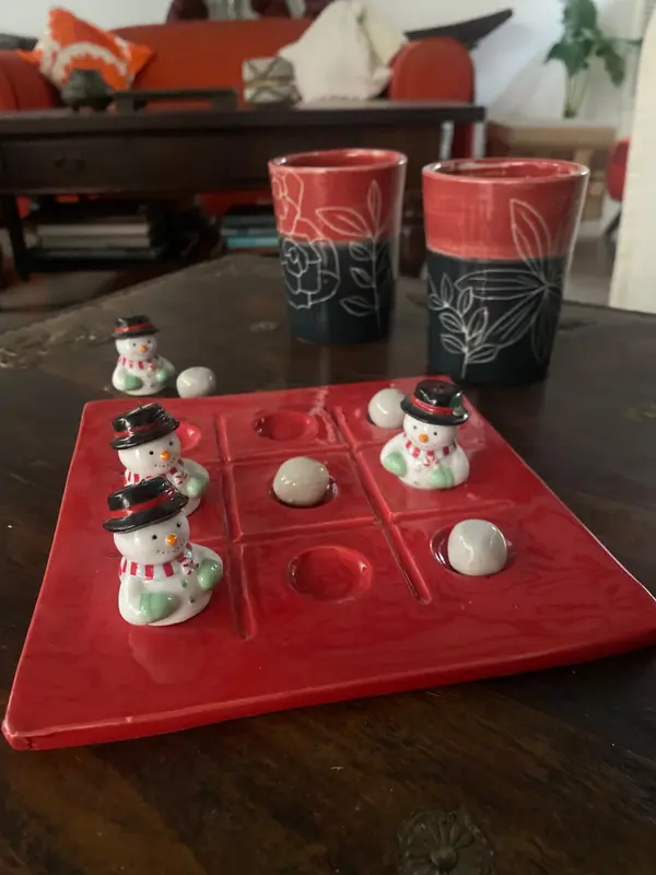 Red ceramic tic-tac-toe board with snowman pieces and two red and black floral mugs on a dark wooden table.