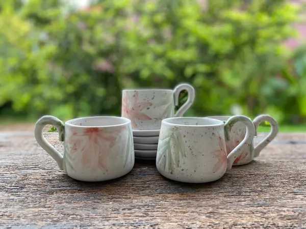 Three white ceramic mugs and saucers with hand-painted pink flowers and green leaves on a wooden table.