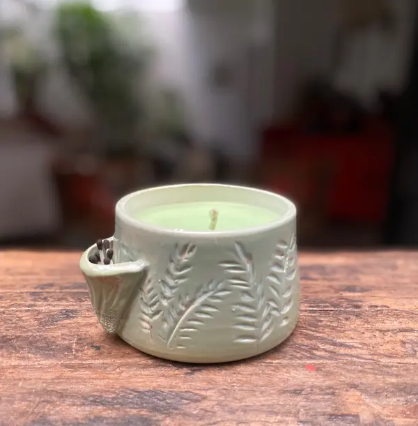 Light green ceramic candle holder with embossed leaf patterns and a side pocket for matches on wood.