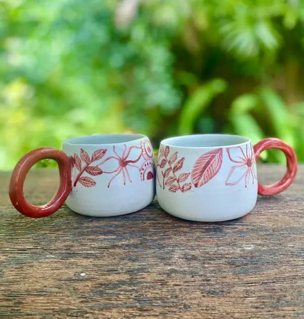 Two white ceramic mugs with red hand-painted floral patterns and large red handles on a wooden surface.