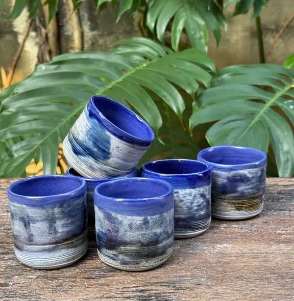 Six small cobalt blue and white glazed ceramic tumblers arranged on a wooden table with tropical foliage behind.