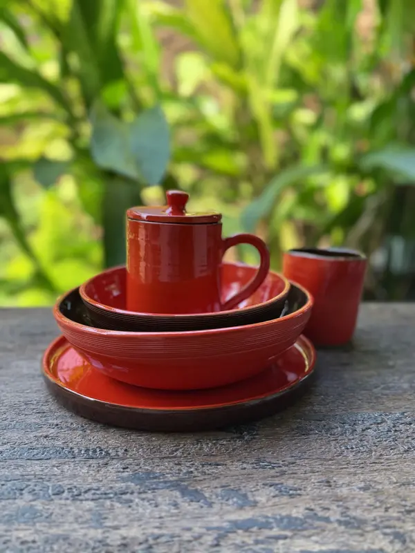 Vibrant red glazed ceramic dinnerware set including a plate, bowls, and lidded mug on a rustic wooden table.