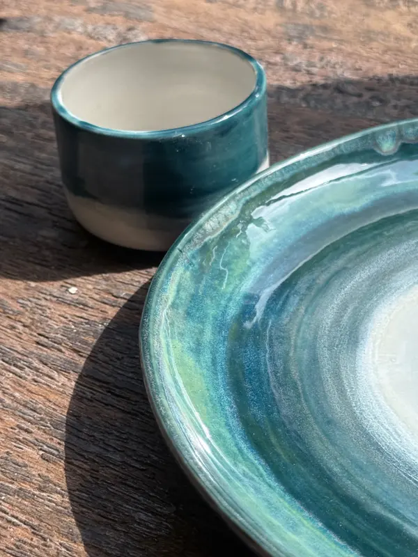 Ceramic plate and small cup with a swirling turquoise and green glaze on a rustic wooden table.