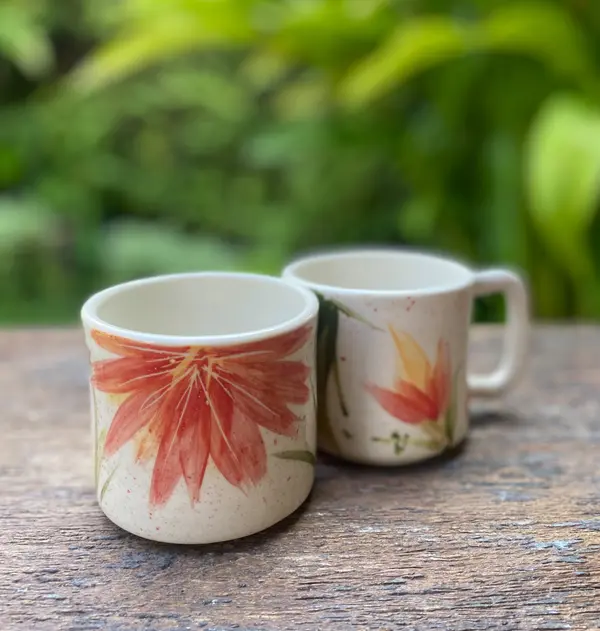Two cream ceramic mugs with hand-painted orange floral designs on a wooden table with a blurred green background.