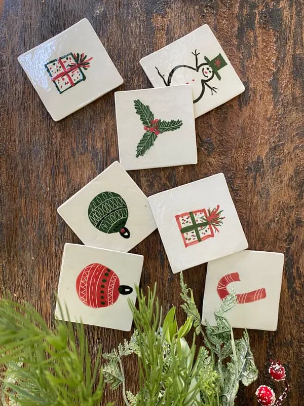Square white ceramic coasters with festive Christmas illustrations on a dark wood surface with greenery.