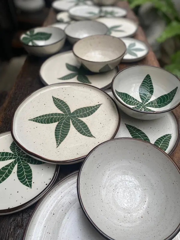 Handmade speckled pottery plates and bowls featuring dark green hand-painted leaf motifs and brown rims.