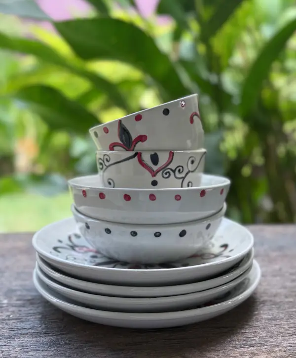 Stack of white ceramic plates and bowls with hand-painted red and black patterns on a wooden table outdoors.