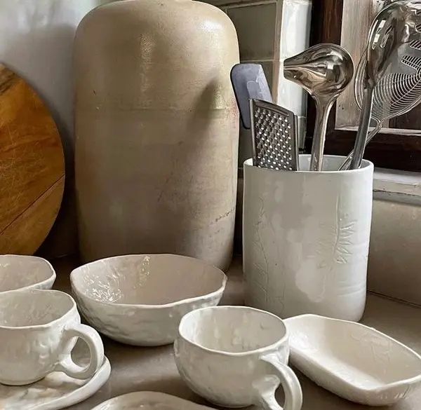 White ceramic kitchenware with organic textures and leaf imprints, including mugs, a bowl, and a utensil holder.