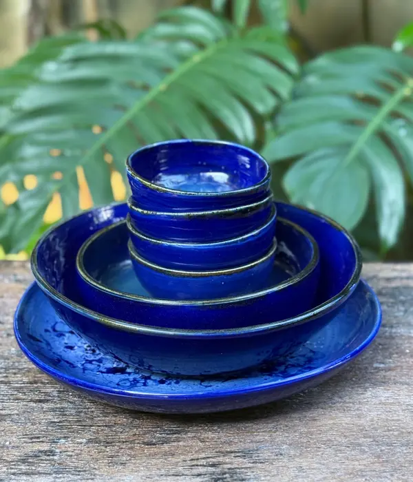 A stack of nested cobalt blue ceramic bowls and a plate on a rustic wooden surface with a leafy background.