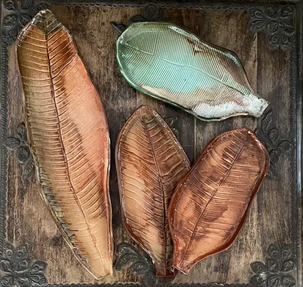 Four ceramic leaf-shaped platters in green and amber glazes with textured veins on a carved wood surface.