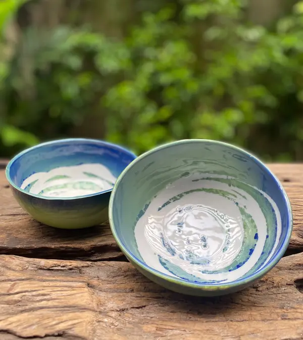 Two ceramic bowls with blue and green swirled glazes on a rustic wood surface with a leafy background.