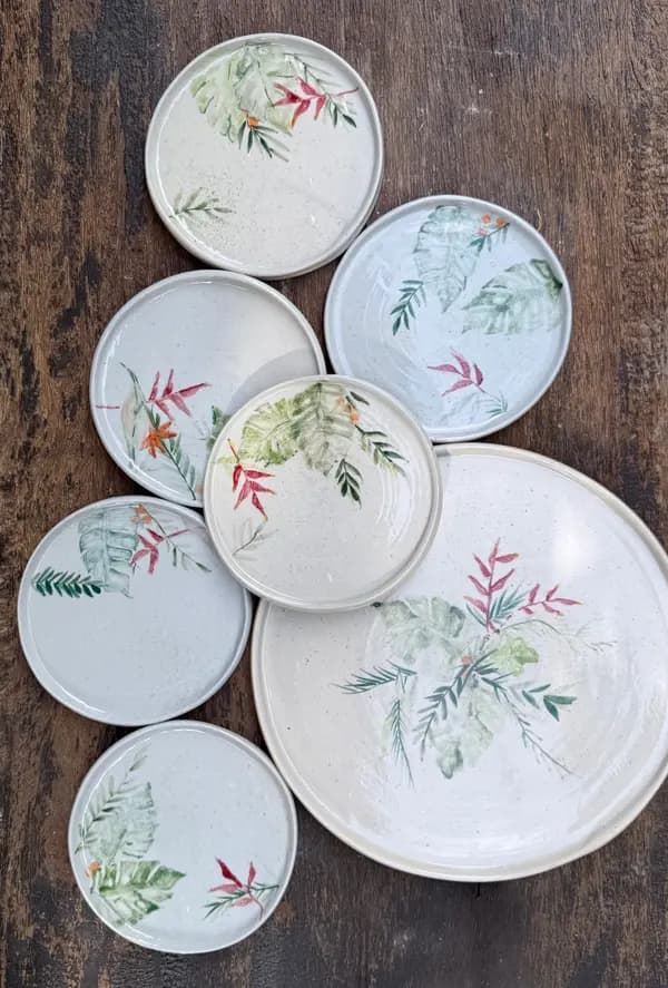White ceramic plates and platter with hand-painted green tropical leaves and red flowers on a wooden surface.