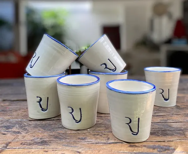 Seven white ceramic cups with cobalt blue trim and hand-painted blue characters on a rustic wood surface.