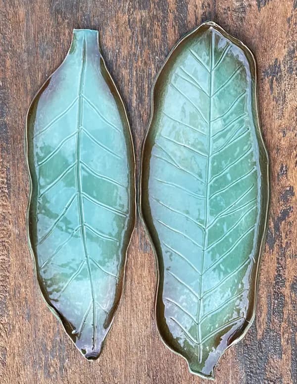 Two ceramic leaf-shaped platters with a turquoise glaze and detailed veins on a dark wood background.