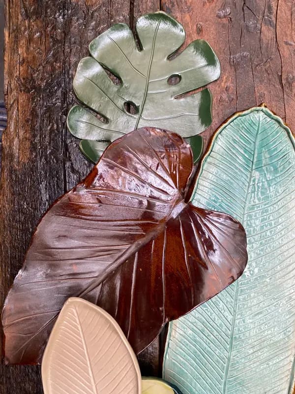 Handmade ceramic platters shaped like Monstera and banana leaves in green, brown, and beige glazes on wood.
