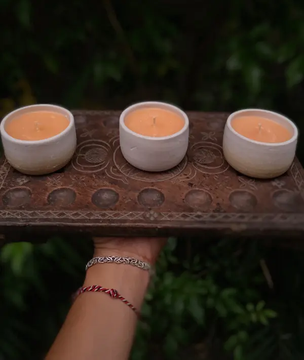 Three small white ceramic candle holders with orange wax on a dark carved wooden board.