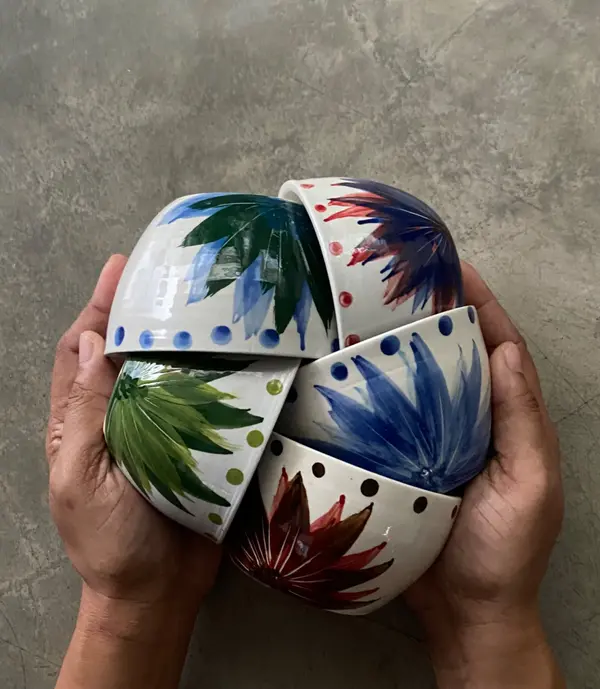 Hands holding five small white ceramic bowls with colorful hand-painted floral designs and dotted rims.