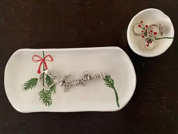 Two white ceramic trinket dishes with hand-painted festive pine and berry motifs, holding jewelry.