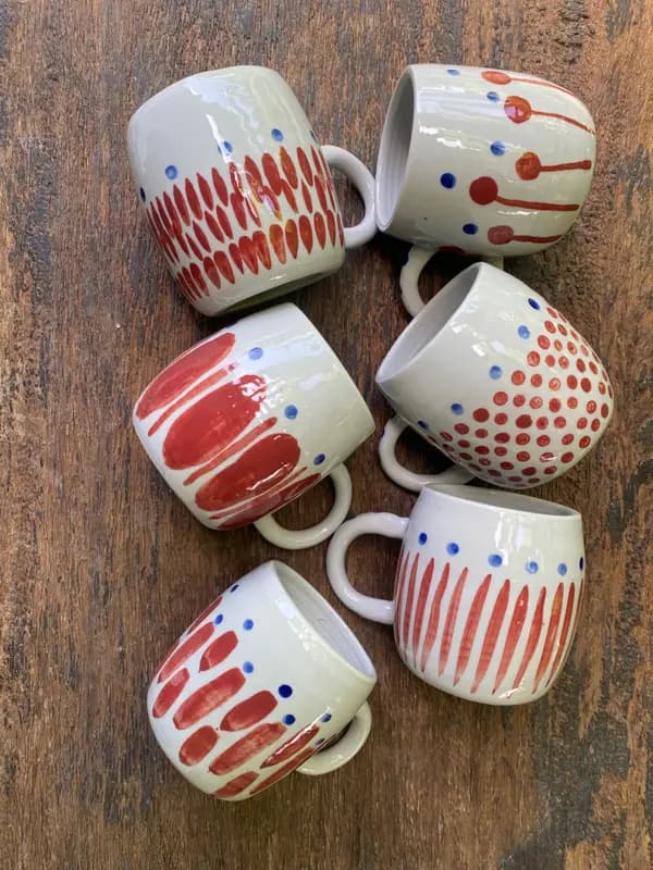 Six white ceramic mugs with red and blue hand-painted patterns on a rustic wooden background.