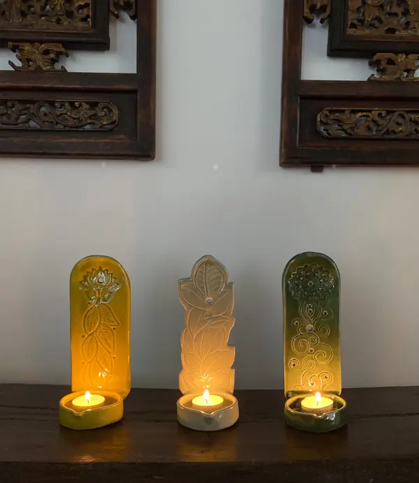 Three vertical ceramic candle holders in yellow, grey, and green with carved botanical patterns and lit tea lights.