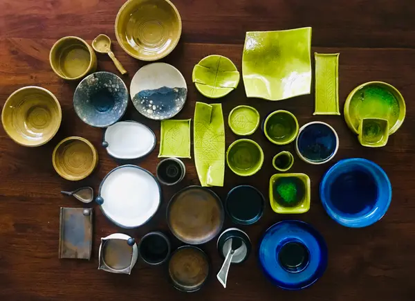 Handmade ceramic bowls, plates, and platters in shades of green, blue, and yellow arranged on a dark wooden surface.