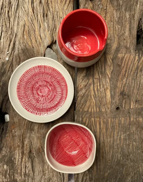 Three red and white ceramic pieces with carved line textures arranged on a rustic, weathered wood surface.
