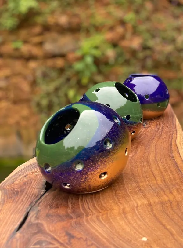 Three spherical ceramic candle holders with green, blue, and orange glazes and perforated holes on a wood slab.