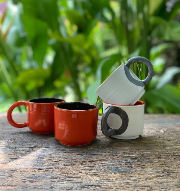 Four handmade ceramic mugs in orange and white with dark handles on a rustic wooden table with garden background.