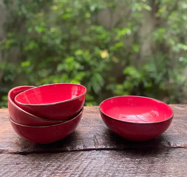 Small glossy red ceramic bowls with dark rims, stacked on a rustic wooden surface with a green garden background.