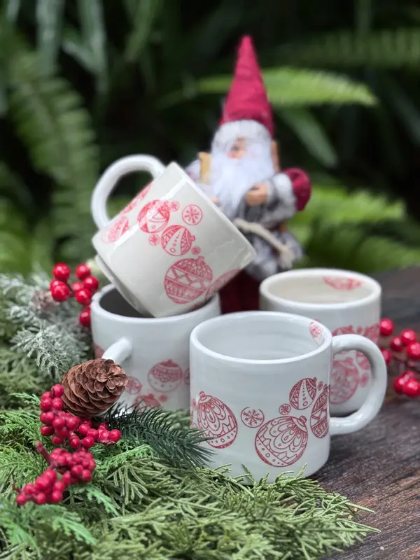 Four white ceramic mugs with red holiday ornament patterns, styled with festive evergreen branches and red berries.