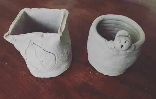 Two unfired white clay pots on a wooden table, featuring leaf impressions and a small sculpted figure on the rim.