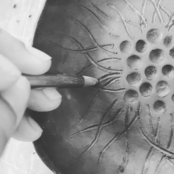 Black and white close-up of a hand carving a floral pattern into a clay bowl with a pointed tool.