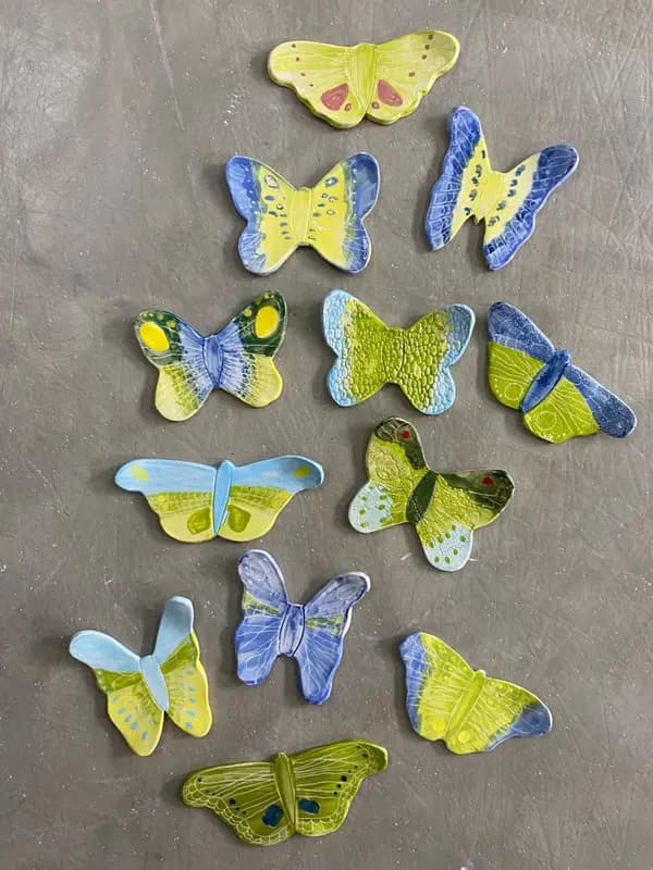 A collection of textured handmade ceramic butterflies in shades of blue, green, and yellow on a grey surface.