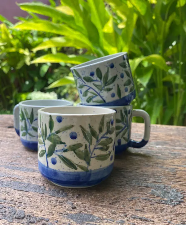 Three hand-painted ceramic mugs with green leaf and blue berry motifs on a white background with a blue base.