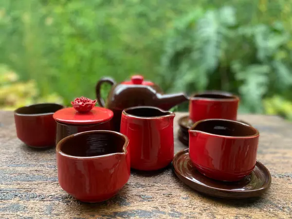 Glossy red and dark brown ceramic tea set with a teapot, cups, and a rose-topped jar on a rustic wooden table.