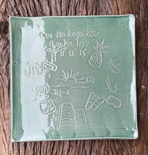 Square sage green ceramic plate with incised tropical illustrations and text on a wooden surface.