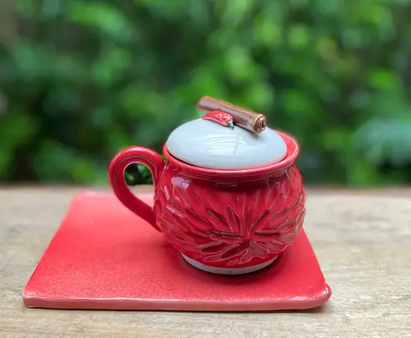 Red textured ceramic mug and square plate set with a white lid featuring a chili and cinnamon stick garnish.