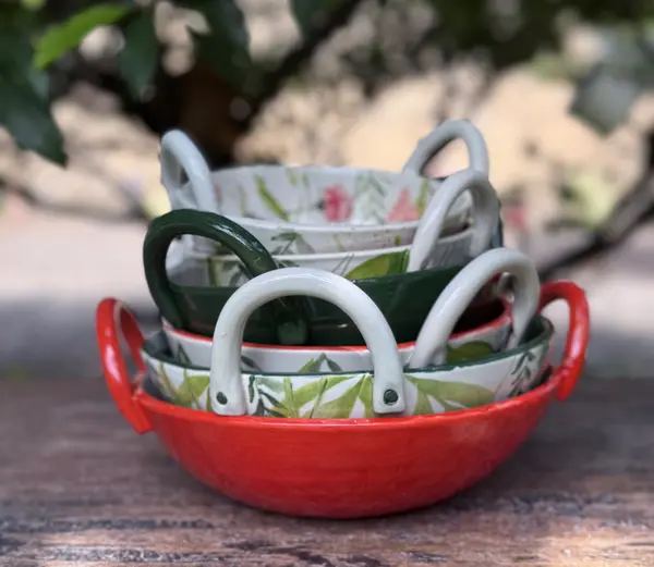 Stack of four nested ceramic bowls with handles, featuring red, dark green, and botanical leaf patterns on white glaze.