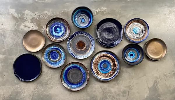 Collection of ceramic plates and bowls with swirled blue, beige, and black glazes on a concrete floor.