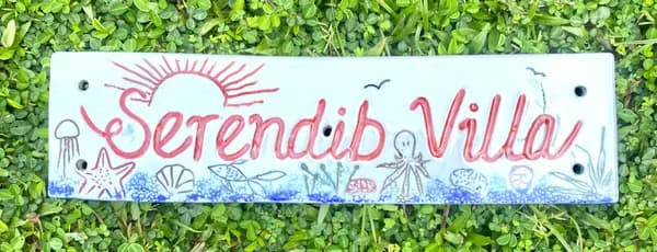 Rectangular ceramic sign for 'Serendib Villa' with red cursive text and hand-painted sea life on a white background.