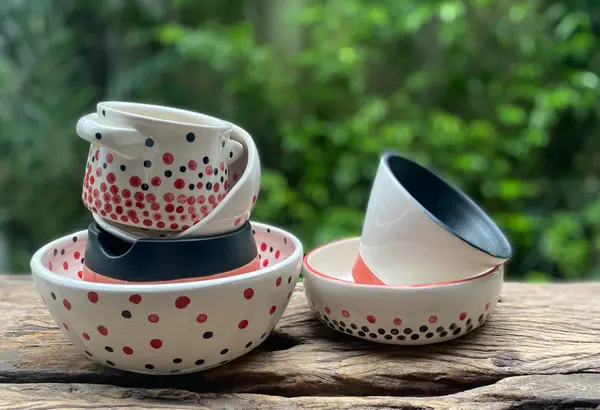 White ceramic bowls and cups with red and black polka dots on a rustic wooden surface with a green blurred background.