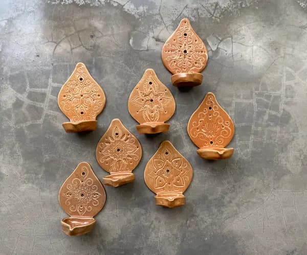 Seven orange terracotta wall-mounted oil lamps with carved mandala patterns on a grey floor.