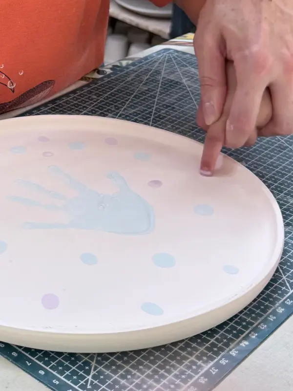 Large beige ceramic plate with a light blue handprint and colored dots, shown during the decorating process.