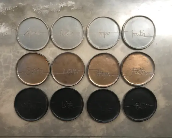Twelve circular ceramic coasters in grey, bronze, and black, inscribed with Spirit, Love, Hope, and Faith.